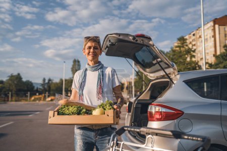Téléchargez les photos : Une femme mature femme caucasienne debout dans le parking bu voiture en face de supermarché épicerie tenant boîte avec de la nourriture et des légumes dans la journée ensoleillée sourire heureux saine manger concept végétalien - en image libre de droit