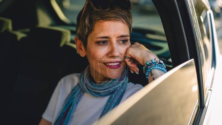 Foto de One mature woman caucasian female sitting on the back seat of the car looking trough the glass window in summer day happy smile travel and transport concept copy space real person gray short hair - Imagen libre de derechos