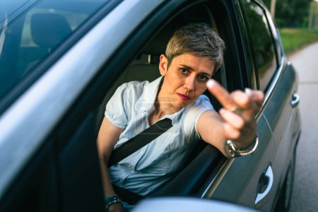 Téléchargez les photos : One woman senior mature female driver showing middle finger trough the window of the car mad angry furious pissed shows a negative gesture to the camera emotionally provocation and rude attitude - en image libre de droit