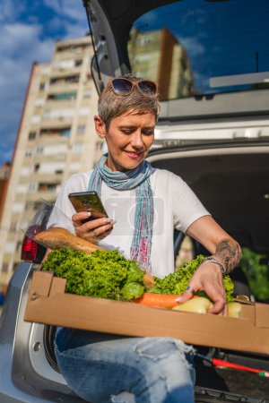 Téléchargez les photos : Une femme mature caucasienne assise dans le coffre arrière de sa voiture sur le parking du supermarché centre commercial ou épicerie avec des légumes nourriture dans la boîte les mettre dans le véhicule - en image libre de droit