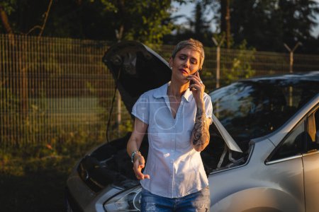Téléchargez les photos : One woman mature female standing on the road in the evening sunset by the broken vehicle car automobile failed engine open hood making a phone call for help roadside assistance towing service concept - en image libre de droit