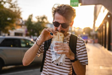 Photo for One man young adult modern caucasian male in the city in sunny day walk and eat sandwich fast food concept urban life copy space tourist eating on the street real person - Royalty Free Image