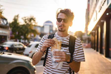Foto de Un hombre joven adulto moderno caucásico masculino en la ciudad en día soleado caminar y comer sándwich concepto de comida rápida vida urbana copiar espacio turístico comer en la calle persona real - Imagen libre de derechos