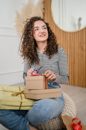Foto de Una mujer joven envuelve regalos en casa feliz sonrisa femenina mientras sostiene papel de regalo y caja de regalo prepararse para empacar regalos para la temporada navideña feliz sonrisa copiar espacio personas reales - Imagen libre de derechos