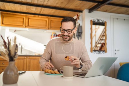 Photo for One man adult caucasian male sit at the table at home having breakfast with laptop computer prepare for work daily morning routine real people copy space - Royalty Free Image