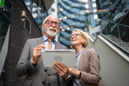 Foto de Senior man dressed in a suit and a mature blonde woman as business partners husband and wife friends or colleagues talk With digital tablet in hand browse internet look data exchange ideas copy space - Imagen libre de derechos