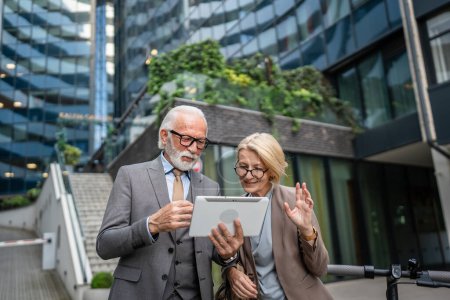 Foto de Senior man dressed in a suit and a mature blonde woman as business partners husband and wife friends or colleagues talk With digital tablet in hand browse internet look data exchange ideas copy space - Imagen libre de derechos