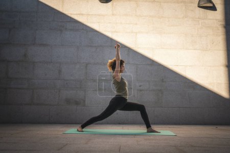 Téléchargez les photos : One adult caucasian woman doing yoga practice in summer day outdoor in the city in front of the wall with shadow and sun healthy lifestyle wellbeing concept wide angle full length self care - en image libre de droit