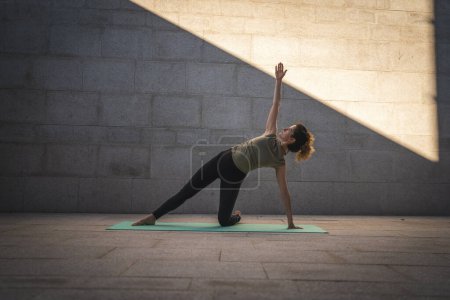 Photo for One adult caucasian woman doing yoga practice in summer day outdoor in the city in front of the wall with shadow and sun healthy lifestyle wellbeing concept wide angle full length self care - Royalty Free Image