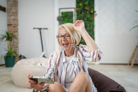 Téléchargez les photos : Une femme caucasienne blonde mature assis intérieur sourire heureux jouer à des jeux vidéo loisirs à la maison ou au travail tenir téléphone mobile utiliser smartphone avoir plaisir à copier en ligne espace porter des lunettes et chemise - en image libre de droit