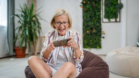 Téléchargez les photos : Une femme caucasienne blonde mature assis intérieur sourire heureux jouer à des jeux vidéo loisirs à la maison ou au travail tenir téléphone mobile utiliser smartphone avoir plaisir à copier en ligne espace porter des lunettes et chemise - en image libre de droit