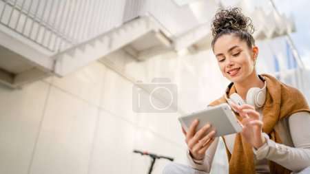 Foto de Joven adulto mujer caucásica estudiante sentarse en el banco al aire libre en el día en frente de la construcción moderna uso tableta digital - Imagen libre de derechos