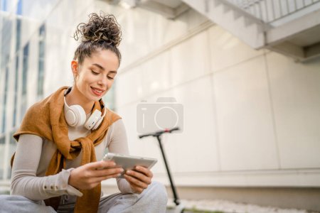 Téléchargez les photos : Jeune adulte femme caucasienne étudiant assis sur le banc en plein air dans la journée devant le bâtiment moderne utiliser tablette numérique - en image libre de droit
