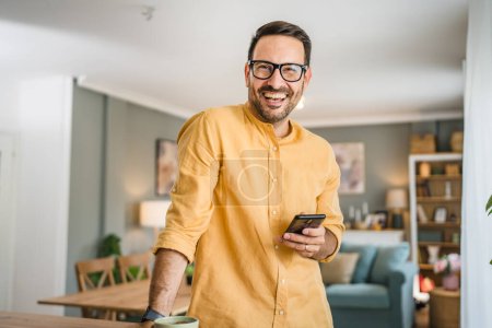 Photo for One man adult caucasian male with beard and eyeglasses stand at home happy smile use mobile phone smartphone to send sms text messages or browse internet online copy space - Royalty Free Image
