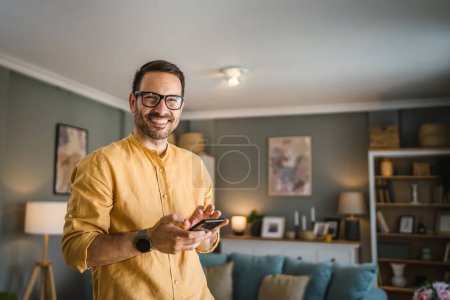 Foto de Un hombre adulto caucásico macho con barba y gafas de pie en casa sonrisa feliz uso de teléfono móvil teléfono inteligente para enviar mensajes de texto SMS o navegar por Internet en línea copiar espacio - Imagen libre de derechos