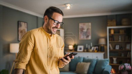 Téléchargez les photos : Un homme adulte caucasien mâle avec barbe et lunettes se tenir à la maison sourire heureux utiliser smartphone téléphone mobile pour envoyer des SMS messages texte ou naviguer sur Internet espace de copie en ligne - en image libre de droit