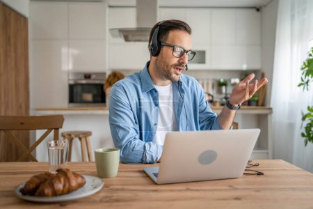 Foto de Un hombre caucásico freelancer masculino trabaja desde casa con auriculares en la cabeza en el ordenador portátil de atención al cliente pensando - Imagen libre de derechos