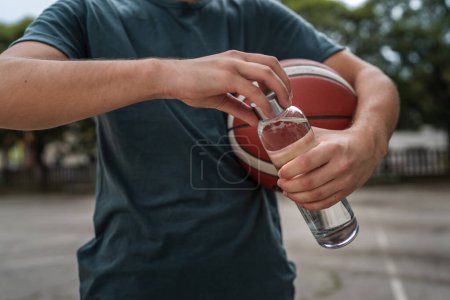 Foto de Primer plano en las manos sección media de hombre caucásico desconocido adolescente abierto botella de plástico de agua al aire libre en día soleado haciendo deportes con baloncesto - Imagen libre de derechos