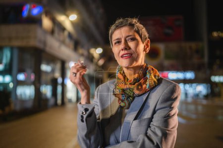 Téléchargez les photos : Une femme âgée caucasienne moderne femme avec des cheveux gris stand dans la ville la nuit ont une cigarette fumer tabac nicotine addiction concept copier espace - en image libre de droit