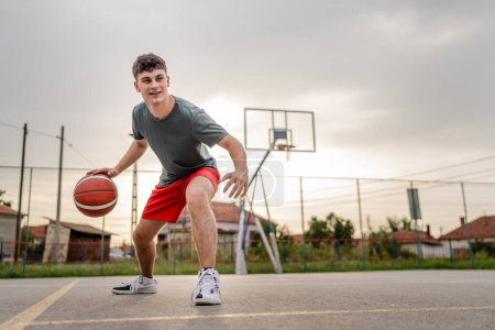 Téléchargez les photos : Un adolescent caucasien mâle caucasien jeune homme debout sur le terrain de basket-ball avec balle dans la soirée prêt à jouer copie jeu espace personne réelle - en image libre de droit