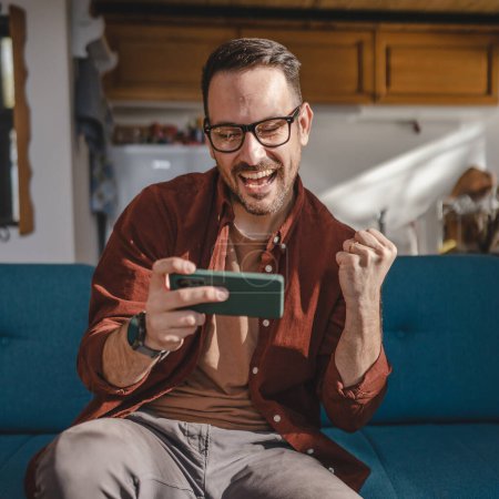 Foto de Un hombre caucásico adulto se sienta en casa feliz sonrisa jugar videojuegos actividad de ocio divertirse celebrar teléfono móvil teléfono inteligente divertirse copiar espacio usar gafas y camisa - Imagen libre de derechos