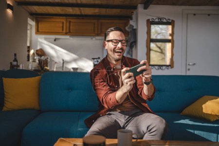 Téléchargez les photos : Un homme caucasien adulte assis à la maison sourire heureux jouer à des jeux vidéo loisirs s'amuser tenir téléphone mobile smartphone avoir plaisir à copier espace porter des lunettes et chemise - en image libre de droit