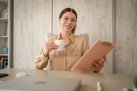 Téléchargez les photos : Une femme jeune adulte caucasienne secrétaire ou gestionnaire ou travailleur assis à la table à son bureau en prenant un frein du travail de vraies personnes copier l'espace heureux sourire tenir tasse de café - en image libre de droit