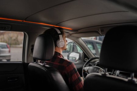 Photo for One man young adult caucasian male sit in the car on the seat with headphones listen guided meditation relaxation practicing mindfulness or listen music or podcast real people copy space back view - Royalty Free Image