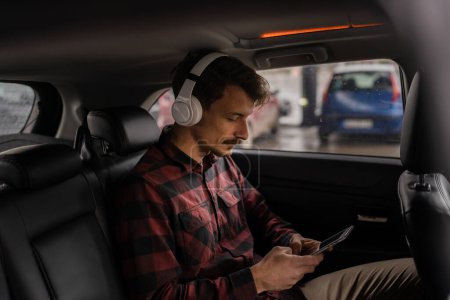 Téléchargez les photos : Un homme adulte caucasien mâle assis dans la voiture avec des écouteurs sur sa tête écouter de la musique ou podcast tout en prenant un frein pendant le voyage d'attente et de détendre les gens réels copier l'espace style de vie moderne - en image libre de droit