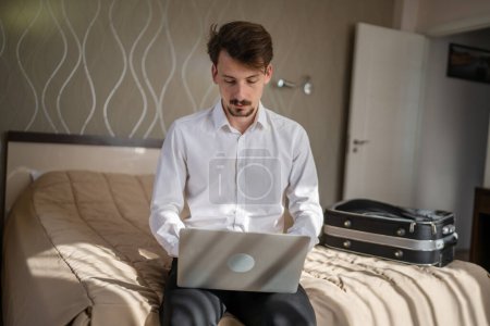 Photo for One man adult caucasian businessman sit on the bed in hotel room work on his laptop computer prepare for meeting or remote online conference real person copy space wear white shirt - Royalty Free Image