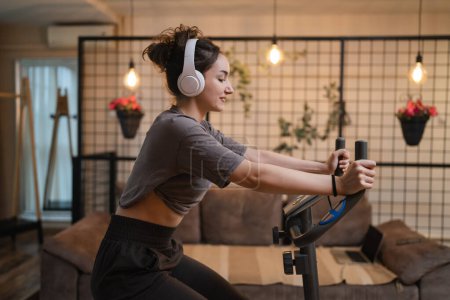 Photo for One woman young caucasian female training at home with headphones on Indoor Cycling stationary Exercise Bike real people copy space health and fitness workout concept side view - Royalty Free Image