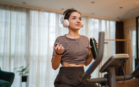 Téléchargez les photos : One woman young caucasian female training at home with headphones on Indoor Cycling stationary Exercise Bike real people copy space health and fitness workout hold bottle of water or supplement - en image libre de droit