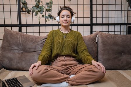 Foto de One woman adult caucasian female using headphones for online guided meditation practicing mindfulness yoga with eyes closed on the sofa bed at home real people self care concept copy space - Imagen libre de derechos
