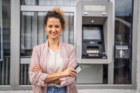 Photo for Portrait of adult caucasian woman stand in front of ATM machine hold credit or debit card - Royalty Free Image