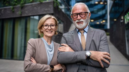 Photo for Portrait of senior man dressed in a suit and a mature blonde woman as business partners husband and wife friends or colleagues stand in front of modern building in day - Royalty Free Image
