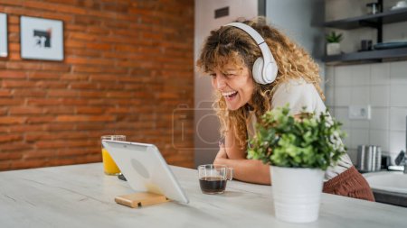 Téléchargez les photos : Une femme caucasienne avec les cheveux bouclés assis à la maison utiliser tablette numérique et écouteurs pour regarder un film ou une série en ligne flux ou d'avoir un appel vidéo sourire heureux avoir un bon espace de copie de temps - en image libre de droit