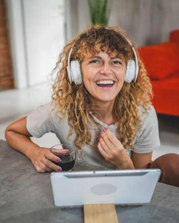 Téléchargez les photos : Une femme caucasienne avec les cheveux bouclés assis à la maison utiliser tablette numérique et écouteurs pour regarder un film ou une série en ligne flux ou d'avoir un appel vidéo sourire heureux avoir un bon espace de copie de temps - en image libre de droit