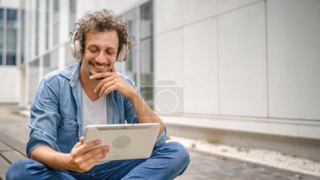 Téléchargez les photos : Un homme caucasien aux cheveux bouclés en plein air devant un bâtiment moderne utilise une tablette numérique et un casque pour regarder un film ou une série en ligne ou pour avoir un espace de copie d'appel vidéo - en image libre de droit