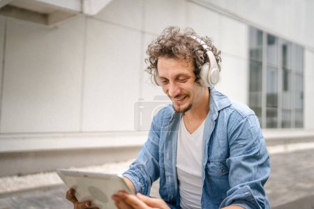Téléchargez les photos : Un homme caucasien aux cheveux bouclés en plein air devant un bâtiment moderne utilise une tablette numérique et un casque pour regarder un film ou une série en ligne ou pour avoir un espace de copie d'appel vidéo - en image libre de droit