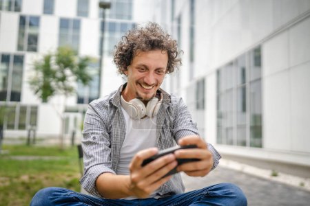 Foto de Un hombre caucásico sentarse al aire libre tomar un freno feliz sonrisa jugar videojuegos actividad de ocio divertirse celebrar el uso del teléfono móvil teléfono inteligente divertirse espacio de copia en línea adulto macho - Imagen libre de derechos