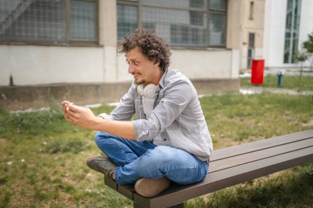 Téléchargez les photos : Un homme caucasien assis à l'extérieur prendre un frein heureux sourire jouer à des jeux vidéo loisirs avoir du plaisir tenir téléphone mobile utiliser smartphone avoir du plaisir copie en ligne espace adulte mâle - en image libre de droit