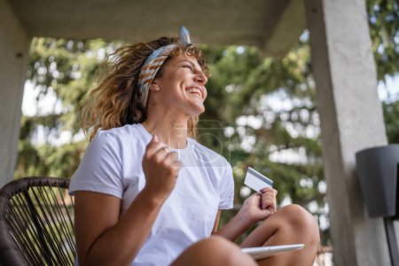 Téléchargez les photos : Une femme faisant des achats en ligne détenir carte de crédit et tablette numérique jeune adulte femme faisant des achats en ligne prendre une décision tout en étant assis sur la terrasse balcon à la maison de vraies personnes copier l'espace sourire heureux - en image libre de droit