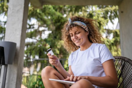 Téléchargez les photos : Une femme faisant des achats en ligne détenir carte de crédit et tablette numérique jeune adulte femme faisant des achats en ligne prendre une décision tout en étant assis sur la terrasse balcon à la maison de vraies personnes copier l'espace sourire heureux - en image libre de droit