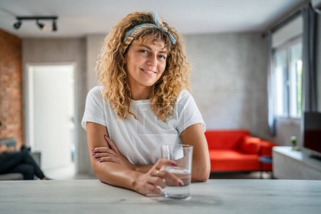 Téléchargez les photos : Une femme caucasienne adulte assis à la maison tenir verre d'eau sourire heureux - en image libre de droit