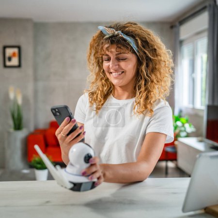 Téléchargez les photos : Une femme caucasienne adulte ajuste et prépare l'application de caméra de surveillance à domicile et utilise un smartphone pour configurer l'espace de copie - en image libre de droit