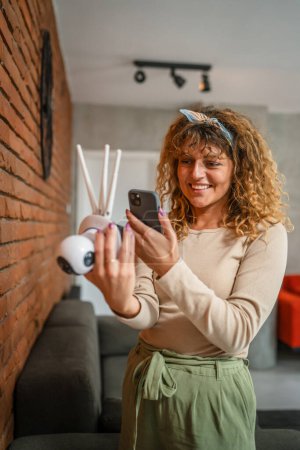 Téléchargez les photos : Une femme caucasienne adulte ajuste et prépare l'application de caméra de surveillance à domicile et utilise un smartphone pour configurer l'espace de copie - en image libre de droit