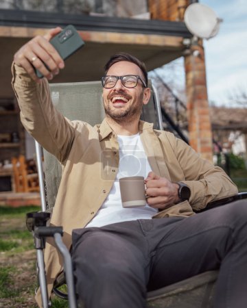 Téléchargez les photos : Un homme heureux caucasien masculin utiliser smartphone pour selfie photo ou vidéo appel extérieur en face de la maison minuscule moderne tout en vacances dans la journée les gens réels copier l'espace - en image libre de droit