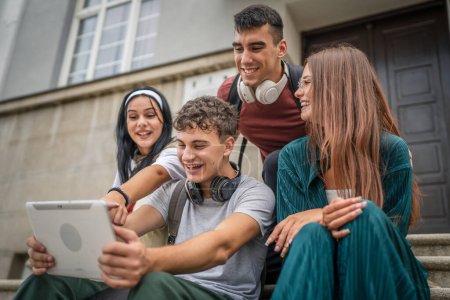 Téléchargez les photos : Groupe d'amis adolescents caucasiens assis en face de l'école ou de l'université détiennent tablette numérique regarder un film vidéo ou avoir un appel vidéo parcourir Internet en ligne pour les personnes réelles de réseau social - en image libre de droit