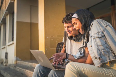 Téléchargez les photos : Adolescent couple caucasien mâle et femelle copain et copine étudiants homme et femme regarder vidéo film sur ordinateur portable à l'école tandis que s'asseoir en plein air dans la journée ensoleillée sourire heureux avoir du plaisir en ligne - en image libre de droit