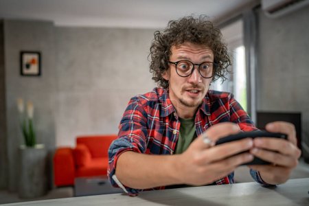 Téléchargez les photos : Homme caucasien assis à la maison jouer à des jeux vidéo sur smartphone téléphone mobile - en image libre de droit